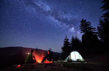 Night-time sojourn in mountains, beneath starry sky. Man and woman, intrepid wanderers, unwind by...