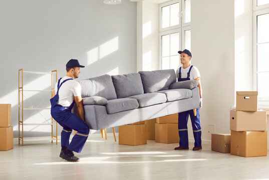 Two young moving service employees workers carrying heavy sofa together in the living room. Men workers from delivery service placing furniture in new house. Moving day and relocation concept.