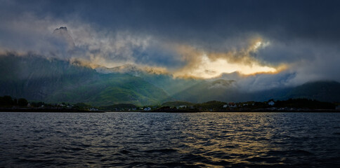 Naklejka na ściany i meble Sun breaking through heavy dark clouds above the mountains and a settlement by the sea. Lofoten Islands, Northern Norway.