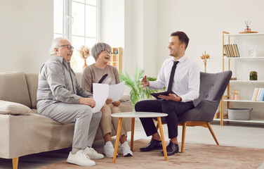 Gray-haired happy smiling senior couple sitting on sofa in the living room at home and having consultation with a male financial advisor about health insurance. Investment in retirement concept - obrazy, fototapety, plakaty