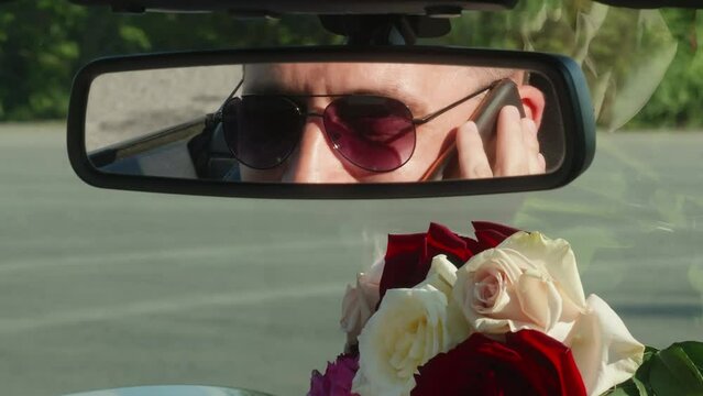 Smart man wearing sunglasses talking on smartphone. Smiling male is reflecting on rear view mirror of car. Closeup of rose bouquet against windshield on dashboard.