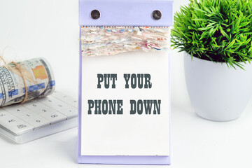 Put Your Phone Down text written on a desktop tear-off calendar on a white background, next to a calculator with a roll of banknotes with a flower out of focus in the background