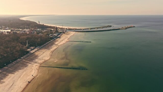 Drone captures sunny Kołobrzeg in February, with tourists strolling, wide sandy beach, calm Baltic Sea, seagulls, lighthouse, and port entrance.