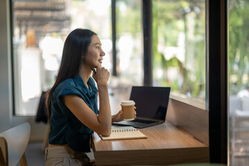 Contemplative young woman looking out window in coffee shop with laptop and coffee cup