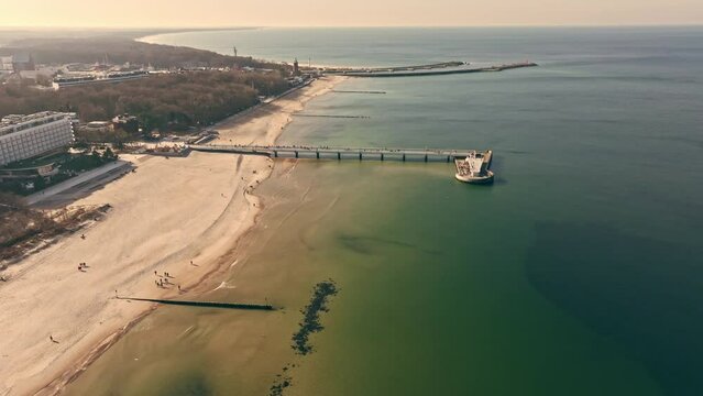 Drone captures sunny Kołobrzeg pier in February, with tourists strolling, wide sandy beach, calm Baltic Sea, seagulls, lighthouse, and port entrance.