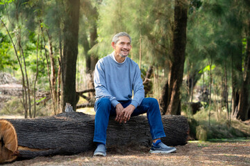 Naklejka na ściany i meble portrait mature man spend leisure time sitting on a log in the forest park,asian senior man smiling and relaxing under a big tree,concept of elderly people lifestyle,hobby,resting,travel