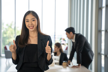 Asian businesswoman gives a thumbs up to the camera with an attractive smile. She stands with team at the office.