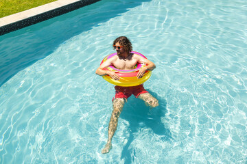 Young Caucasian man floats in a pool with a colorful ring, wearing sunglasses and red shorts