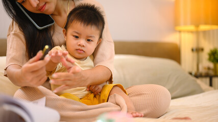 Cute little baby boy sitting on the bed while busy mother working and talking on mobile phone