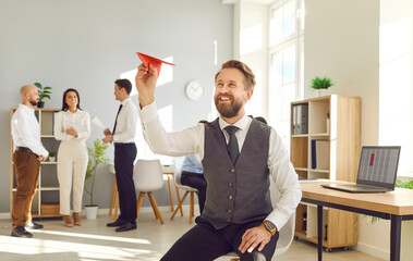 Happy male leader of company throwing red paper plane in office with group of employees smiling and...