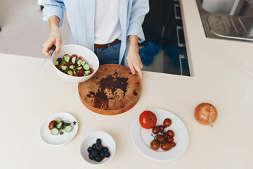 Woman preparing healthy salad bowl with fresh ingredients on wooden cutting board, surrounded by other bowls of food