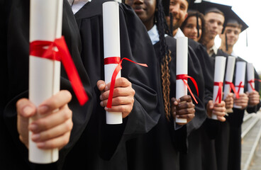 Close up hands of diverse graduate students holding diplomas and standing in a row in black robes outdoor at graduation ceremony. Education and graduating from university or college concept.