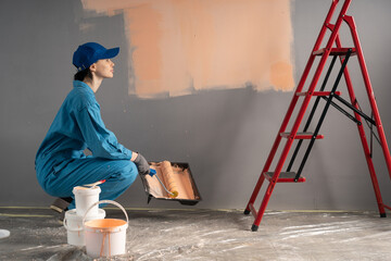 A painter in a cap is squatting near a wall, ready to paint the walls with a paint roller. Home...