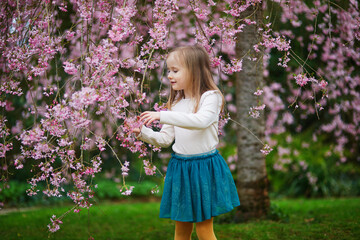 Adorable preschooler girl enjoying nice spring day in park during cherry blossom season