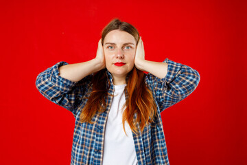 Young beautiful 30s woman covering her ears while standing against red studio background. The concept hear no evil