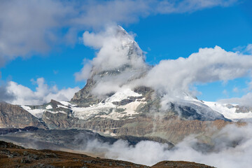 Fototapeta na wymiar Low angle view of near symmetric pyramidal peak of the Matterhorn near Zermatt, Switzerland in extended Monte Rosa area of the Pennine Alps somewhat surrounded by white clouds in otherwise blue sky