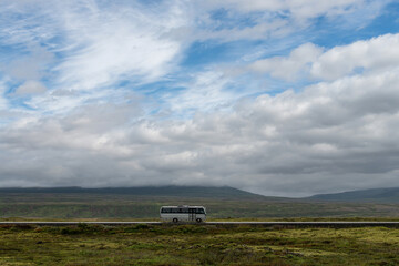 Side view of a bus on the only highway or ring road (&THORN;j&oacute;&eth;vegur) of Iceland on an otherwise empty road against the backdrop of the empty landscape of green mountains partly covered in thick clouds