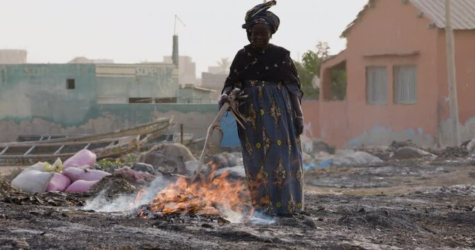 Close-up. Elderly Black African woman smoking small fish on wood chips. Air pollution. Dakar, Senegal