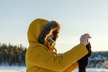 Woman in yellow winter jacket taking a photo with her smart phone at sunset in Lapland