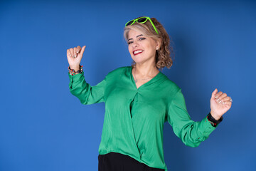 Pretty cheerful woman in green blouse and green glasses on a blue background dancing, Image with copy space for text.