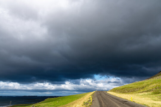 Low angle view of a sun lit gravel road and green pasture in the northern part of Iceland with above that large dark rain clouds giving a very dramatic look to the landscape