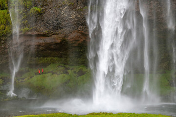 Close up the Seljalandsfoss waterfall in Iceland with some people in red rian coats walking in spray and mist on footpath behind the waterfall with its origin in the volcano glacier Eyjafjallajokull