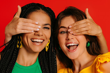 Two playful young women in colorful wear covering eyes to each other and smiling against red background - 749384016