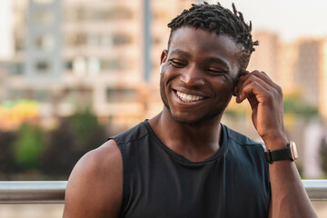 Cheerful African man in sportswear adjusting his headphones while resting after training outdoors - 749385495