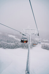 Tourists in colorful ski suits ride on a chairlift over a snowy forest uphill