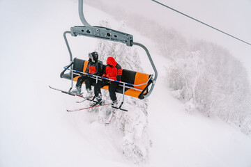 Skiers ride a chairlift up a foggy mountain above a snowy forest