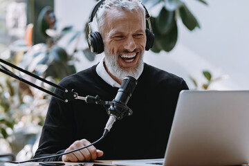 Happy mature podcaster in headphones using microphone while broadcasting from studio - 749397871