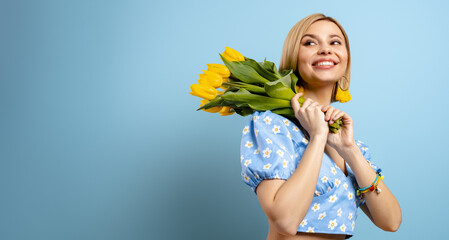 Happy young woman carrying bunch of yellow tulips on shoulder and against blue background - 749403464