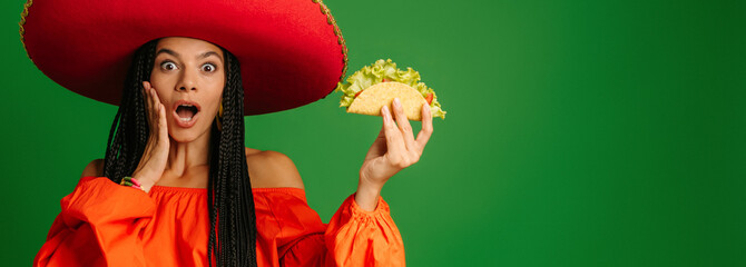 Attractive young Mexican woman in Sombrero holding taco and looking surprised against green background - 749417058