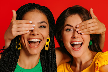 Two happy young women in colorful wear covering eyes to each other and smiling against red background - 749417273