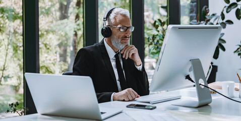 Confident mature businessman in headphones using computer while having web conference from office - 749417608