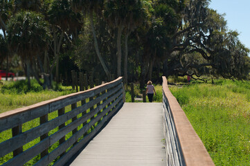 Leading lines down the nature trail wood bridge with a woman at the end walking away. Wide view with grass on  the sides, sky and trees in the background. At Myakka River State Park in Sarasota FL