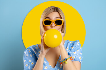 Playful young woman blowing up a balloon while standing against blue background - 749420696