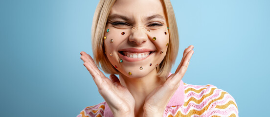 Playful young woman with colorful rhinestones over her face standing against blue background - 749421210