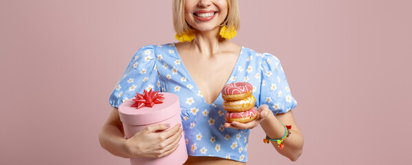 Close-up of surprised young woman holding gift box and doughnuts on pink background - 749422027