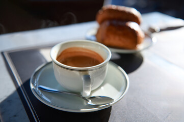 Elegant white ceramic cup of freshly brewed espresso coffee with foam on saucer. Blurred crispy croissants on background