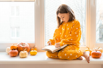Cute little girl in an orange costume sits with decorative pumpkins on a window sealing. Halloween concept. Fall themed photo of a child with pumpkins. Kid with autumn season indoor decorations.
