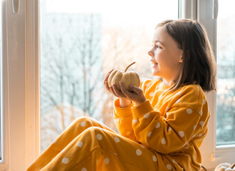 Cute little girl in an orange costume sits with decorative pumpkins on a window sealing. Halloween...