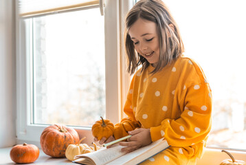 Cute little girl in an orange costume sits with decorative pumpkins on a window sealing. Halloween concept. Fall themed photo of a child with pumpkins. Kid with autumn season indoor decorations.