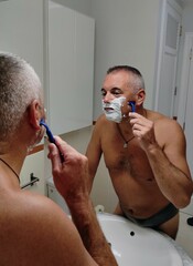 Middle-aged man, with short, graying hair, bare-chested and wearing underwear, shaving in front of...