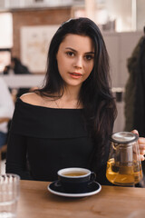 Brunette woman sitting at table drinking tea in restaurant. Teen woman wear black long sleeves top hold teapot with green tea, cups in cafe. Happy girl relaxing and rest with friends.