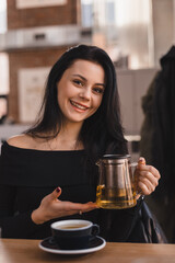 Brunette woman sitting at table drinking tea in restaurant. Teen woman wear black long sleeves top hold teapot with green tea, cups in cafe. Happy girl relaxing and rest with friends.