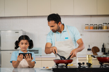 curious father with daughter cooking together by watching online video from mobile phone at kitchen - concept of family bonding, weekend activities and teamwork