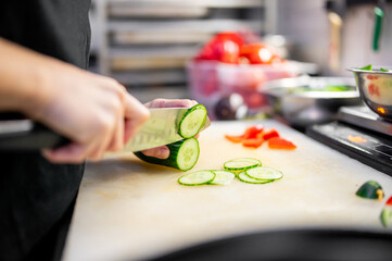 A chef deftly slices fresh cucumber on a cutting board in a professional kitchen. Chopped...