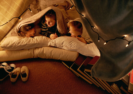 Kids, happy and sleepover in tent at night with conversation, bonding and holiday adventure or vacation. Funny friends or children by fairy lights, pillows and blanket at home for fun storytelling
