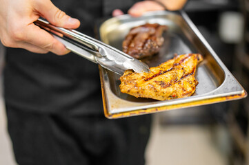 Close-up of a chef holding a tray with grilled chicken and steak using tongs. The chef, dressed in black, serves the deliciously grilled food.